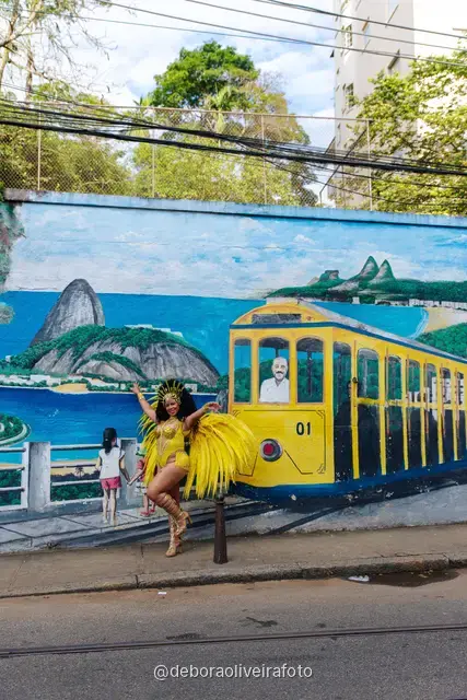 Mujer con disfraz de Carnaval amarillo frente a un mural con un tranvía amarillo y el Pan de Azúcar en Santa Teresa, Río de Janeiro