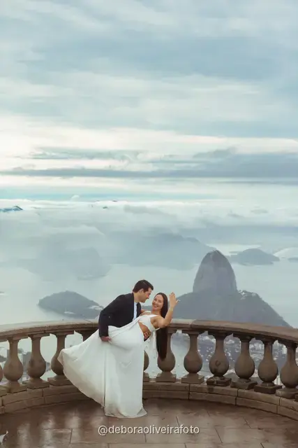 Couple showing a ring and embracing at Christ the Redeemer, with Sugarloaf Mountain in the clouds behind