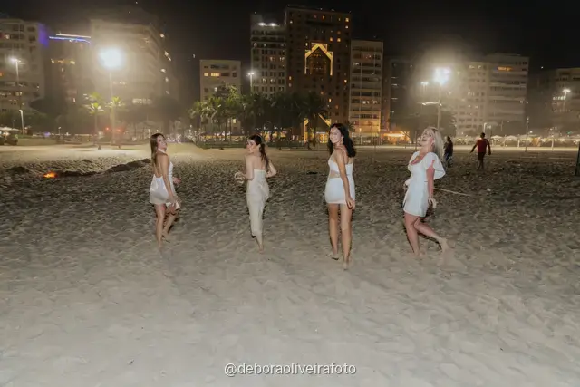Group of four women in white posing on the sand at Copacabana with the city lights behind at night