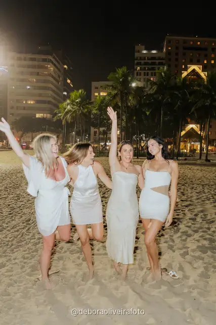 Grupo de quatro mulheres de branco posando na areia de Copacabana com as luzes da cidade atrás, à noite