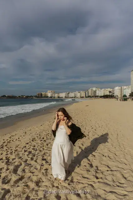 Woman in a white lace dress and green jacket sitting on the sand of Copacabana Beach