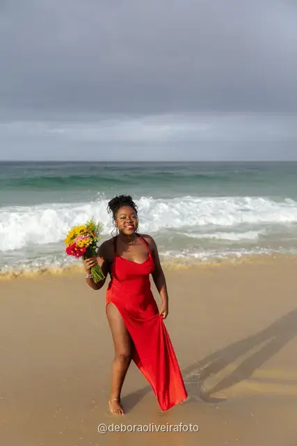 Woman in a long red dress holding a bouquet of sunflowers at the water line of Ipanema Beach in Rio de Janeiro