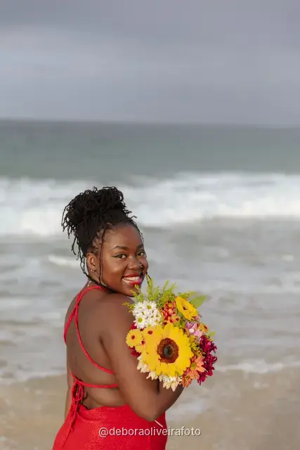 Woman in a long red dress holding a bouquet of sunflowers at the water line of Ipanema Beach in Rio de Janeiro