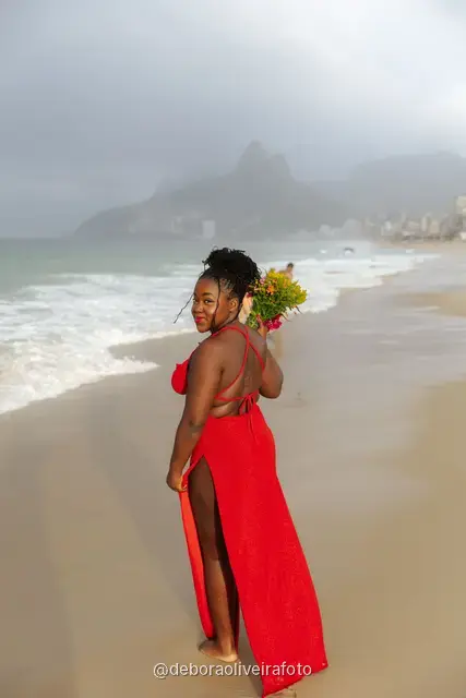 Woman in a long red dress holding a bouquet of sunflowers at the water line of Ipanema Beach in Rio de Janeiro