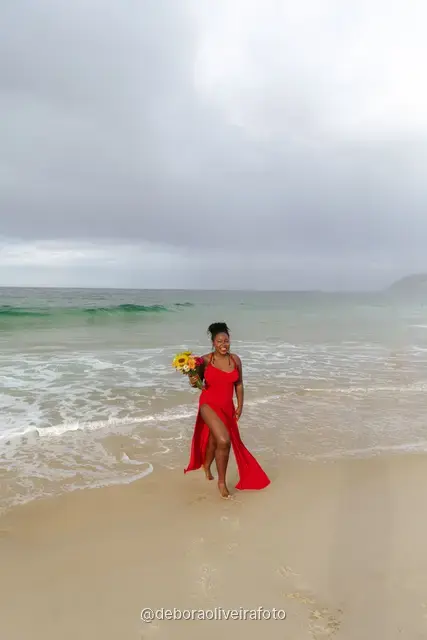 Woman in a long red dress holding a bouquet of sunflowers at the water line of Ipanema Beach in Rio de Janeiro