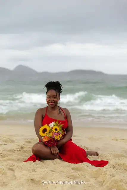 Woman in a red dress sitting on the sand of Ipanema Beach with a sunflower bouquet